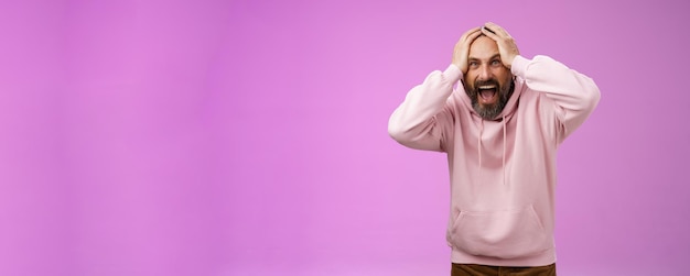 Free photo outraged furious mad adult man bearded yelling shocked despair hatefully looking camera insane holding hands head frowning shouting losing bet disappointed very upset standing purple background