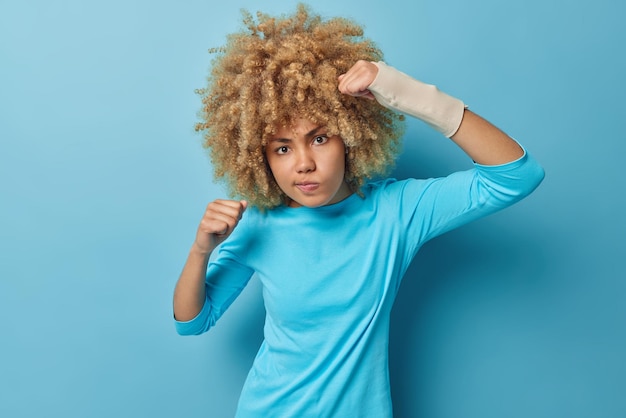 Free photo outraged irritated woman with curly hair clenches fists angrily frowns face wears elastic band on arm after accident got injured dressed casually isolated over blue background insurance concept