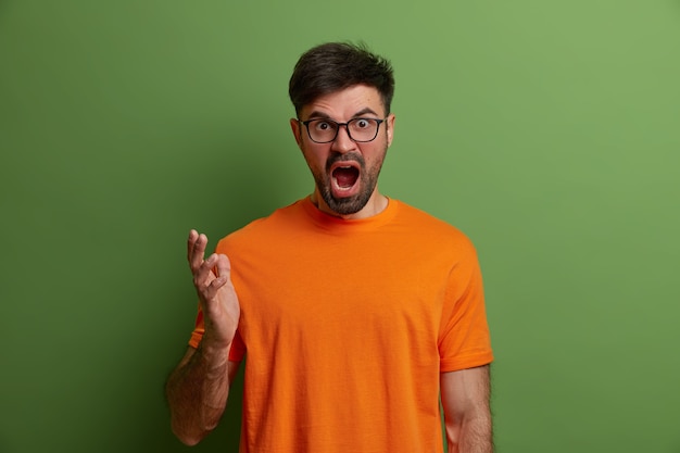 Free photo outraged stressful european man screams from annoyance, gestures angrily, argues with someone, looses temper and feels aggression, shakes hands, wears orange t shirt, isolated on green wall