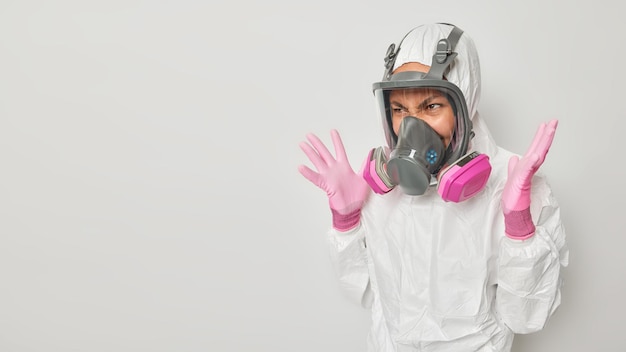 Free photo outraged woman dressed in protective suit with hood wears gas mask keeps palms raised realizes being under threat in difficult situation isolated over white background blank space for promo