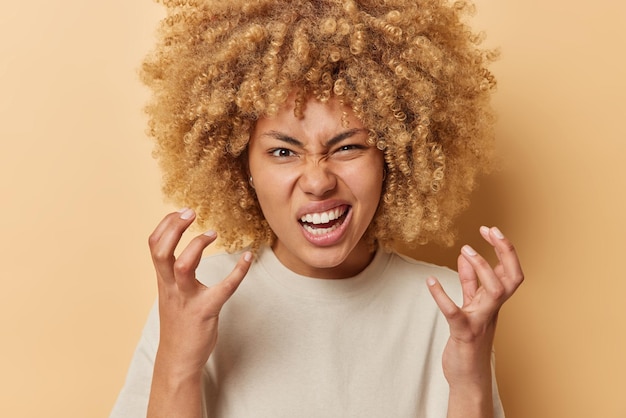 Free photo outraged young woman with curly bushy hair gestures angrily frowns face expresses negative emotions dressed in casual t shirt isolated over beige background grimaces at something ridiculous