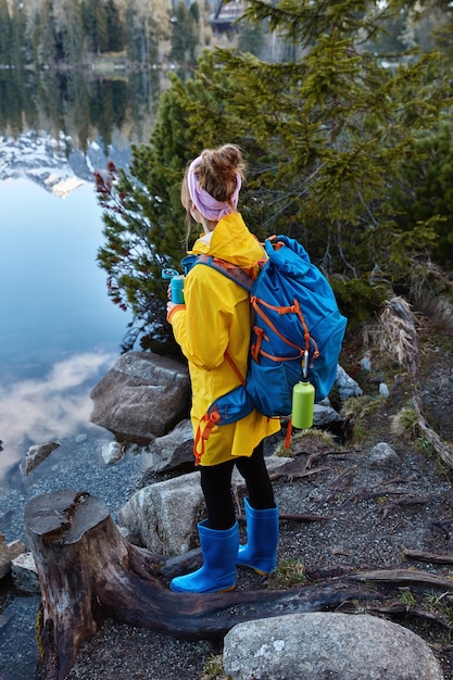 Outside shot of female traveler enjoys panoramic mountain lake, drinks hot tea during rest after strolling, carries big rucksack, has holiday trip