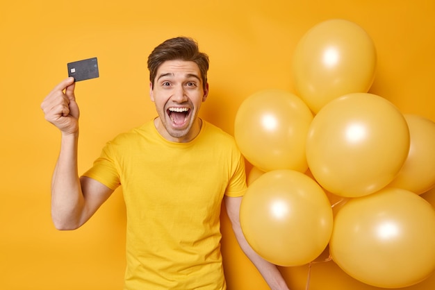 Free photo overemotive cheerful young man in casual t shirt holds credit card and bunch of helium balloons ready for celebration going to spend all money isolated ove yellow background celebrates birthday