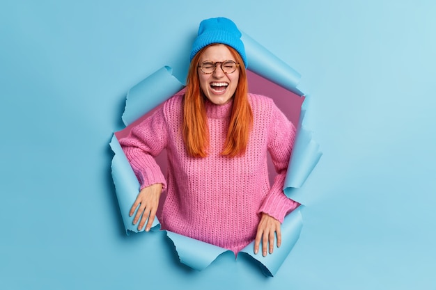 Overemotive ginger young European woman smiles joyfully and exclaims from happiness dressed in casual wear wears optical glasses stands indoor in paper torn hole