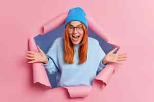 Free photo overemotive positive ginger young woman exclaims loudly spreads palms feels very glad wears blue hat and jumper breaks through pink paper