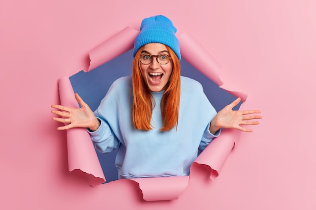 Free Photo overemotive positive ginger young woman exclaims loudly spreads palms feels very glad wears blue hat and jumper breaks through pink paper