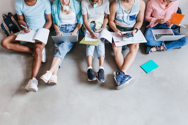 Free photo overhead portrait of students in trendy sneakers chilling on the floor while preparing for exams together. university friends spending time together using laptops and writing abstract.