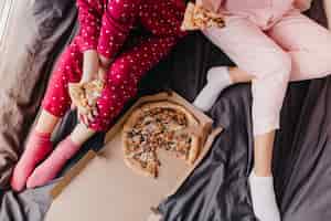 Free photo overhead portrait of two girls in pajamas sitting on bed with italian fast food. lazy female models eating pizza on dark sheet.