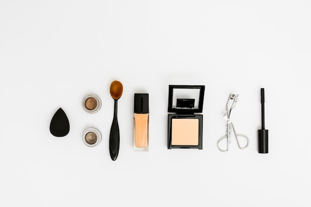 An overhead view of blender; eyeshadow; oval brush; compact powder; eyelash curler and mascara brush on white backdrop