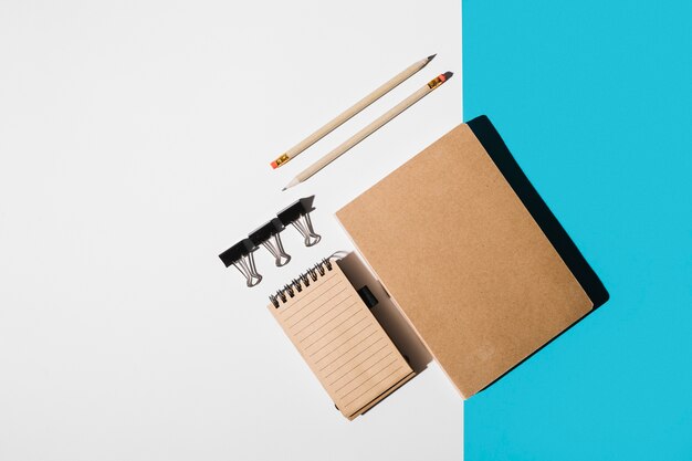 An overhead view of book; spiral notepad; pencil; bulldog clips on white and blue backdrop