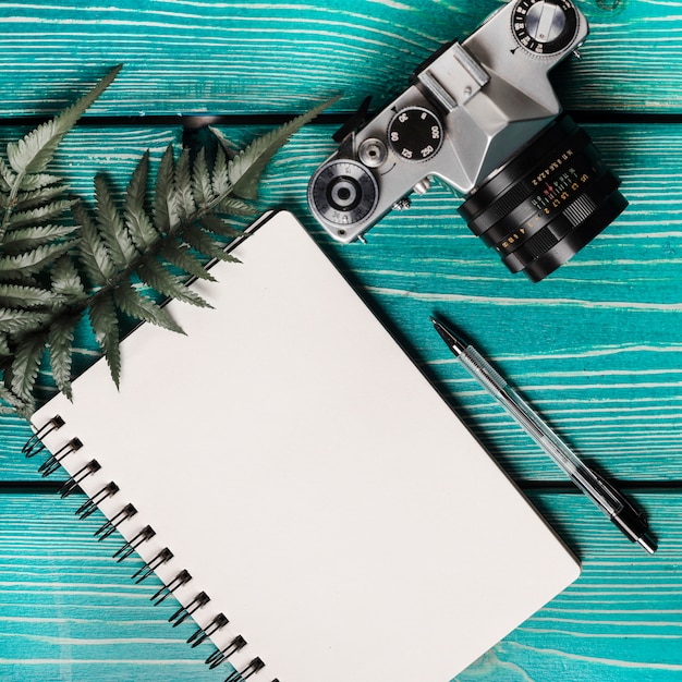 Free photo an overhead view of camera; fern; pen and blank spiral notepad on wooden textured background