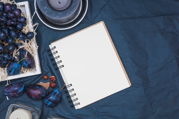 Free photo an overhead view of candles; blank spiral notepad; plums and grapes on textile backdrop