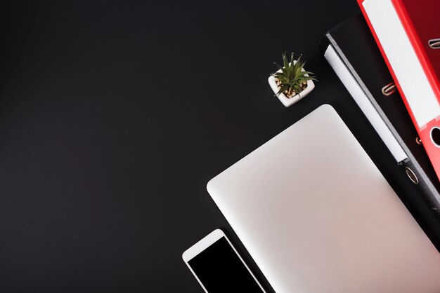 An overhead view of cell phone; laptop; cactus plant and files on black background