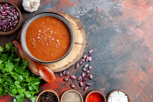 Free photo overhead view of classic tomato soup in a blue bowl spoon on wooden tray garlic and a bunch of green on mixed color table