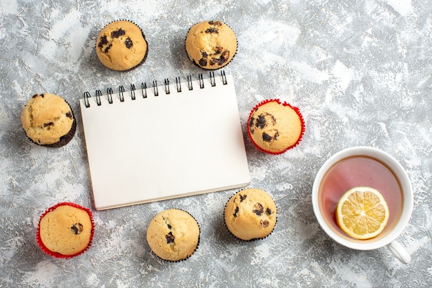 Free photo overhead view of closed notebook among delicious small cupcakes with chocolate and hand holding a cup of black tea with lemon on ice surface