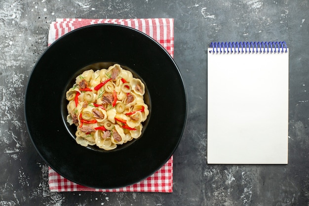 Free photo overhead view of delicious conchiglie with vegetables and greens on a plate and knife on red stripped towel and notebook on gray background