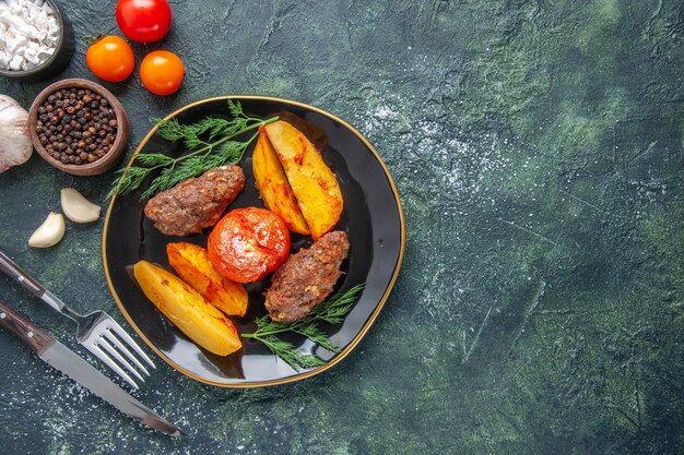 Overhead view of delicious meat cutlets baked with potatoes and tomatoes on a black plate cutlery set spices garlics tomatoes on the right side