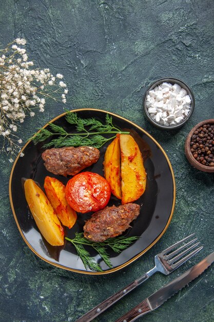 Overhead view of delicious meat cutlets baked with potatoes and tomatoes on a black plate cutlery set white flowers spices on green black mixed colors background