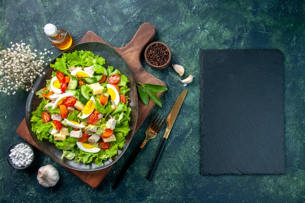 Overhead view of delicious salad with fresh ingredients on wooden cutting board spices oil bottle garlics cutlery set on black mix colors background