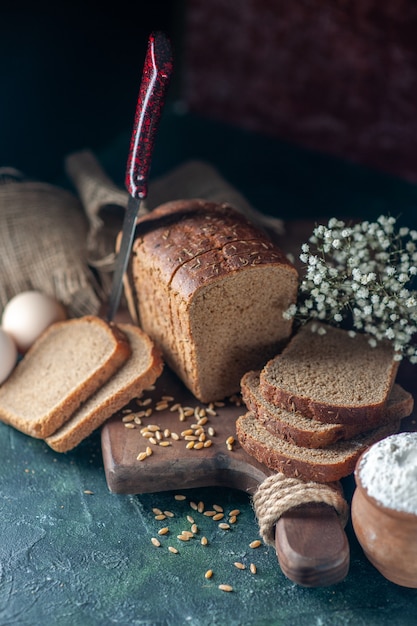 Overhead view of dietary black bread wheats on wooden cutting board knife flower eggs flour in bowl brown towel on mixed colors background