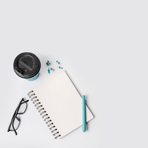 Free photo an overhead view of disposable coffee cup; push pins; pen; eyeglasses and spiral notepad on white background