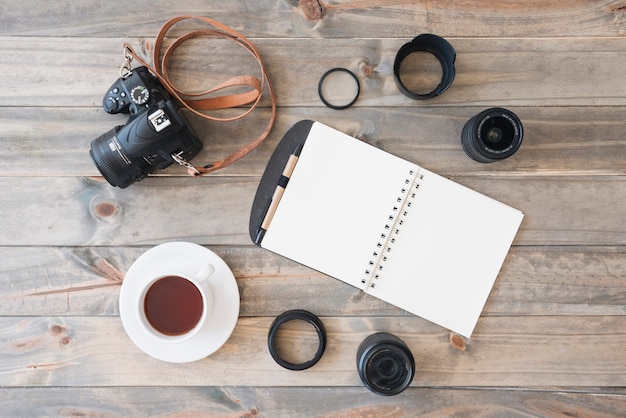 Free photo overhead view of dslr camera; cup of tea; spiral notepad; pen; camera lens and extension rings on wooden background