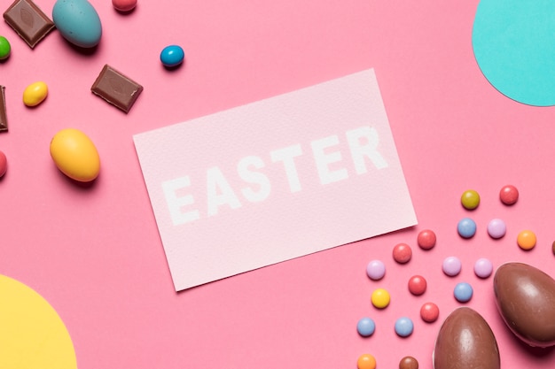 An overhead view of easter word with chocolate easter eggs and gem candies on pink background