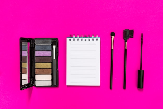 An overhead view of eyeshadow palette; spiral notepad and makeup brushes on pink background