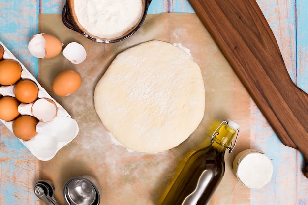 An overhead view of flat dough; flour; eggs; measuring spoon and oil on wooden table