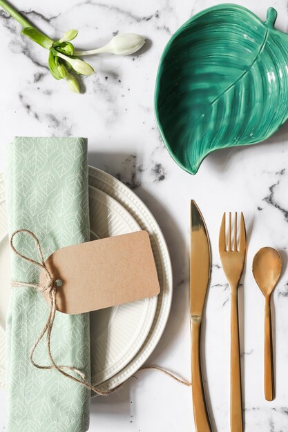 An overhead view of folded napkin tied with tag on ceramic plate and cutlery over marble textured background