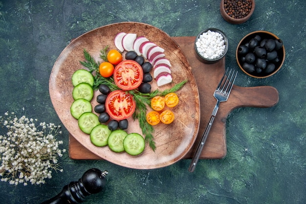 Free photo overhead view of fresh chopped vegetables in a brown plate on wooden cutting board olives in bowl salt garlics flower on mixed colors background