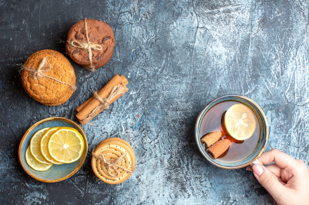 Overhead view of fresh lemons and hand holding a cup of black tea with cinnamon various stacked cookies on dark background