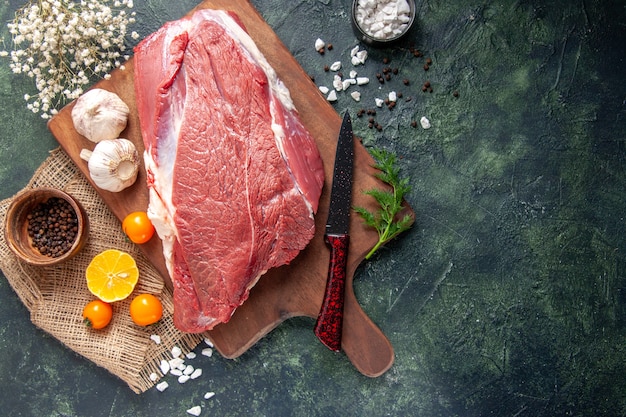 Overhead view of fresh raw red meats green garlics on brown wooden cutting board tomatoes pepper on nude color towel knife on dark color background