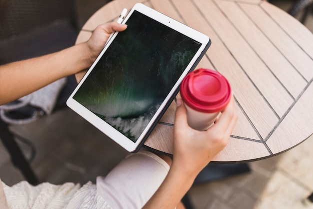 An overhead view of a girl holding digital tablet and takeaway coffee cup on table