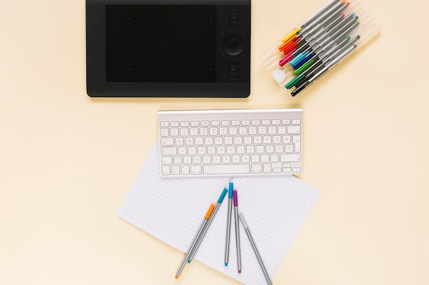 Overhead view of graphic digital tablet with keyboard and felt-tip pens on notebook over the beige background