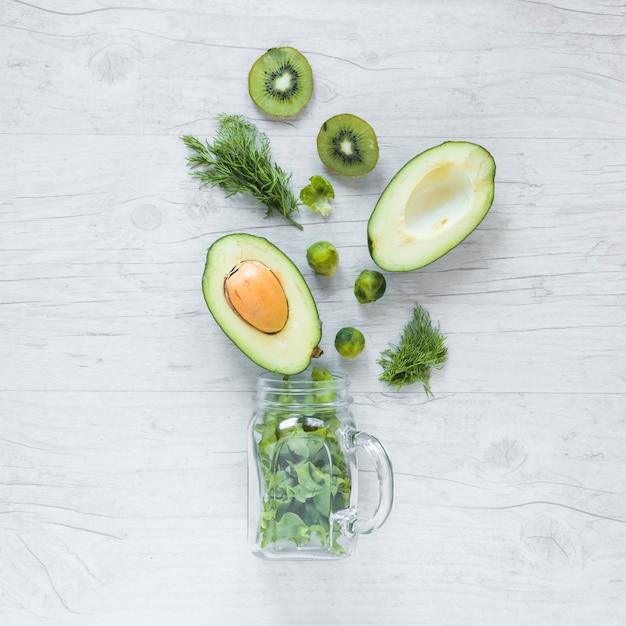 Free photo overhead view of halved avocado; kiwi; brussels sprouts and lettuce with jar against wooden background