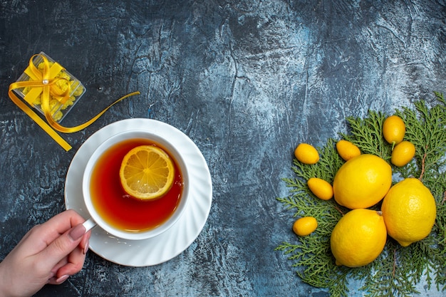 Overhead view of hand holding a cup of black tea with lemon and yellow gift box next to citrus fruits collection on fir branches on dark background