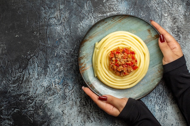 Overhead view of hand holding delicious pasta meal on a blue plate served with tomato and meat for dinner on the left side on ice background