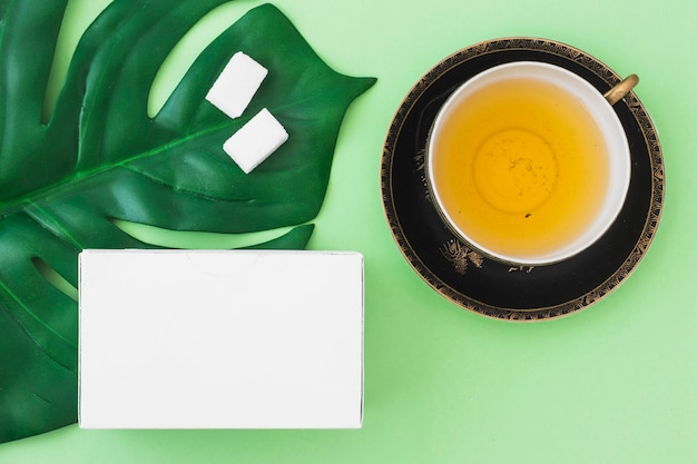 Overhead view of herbal tea cup with sugar cubes and white box on green background