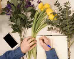 Free photo an overhead view of a male florist writing on diary with pen on table with flowers