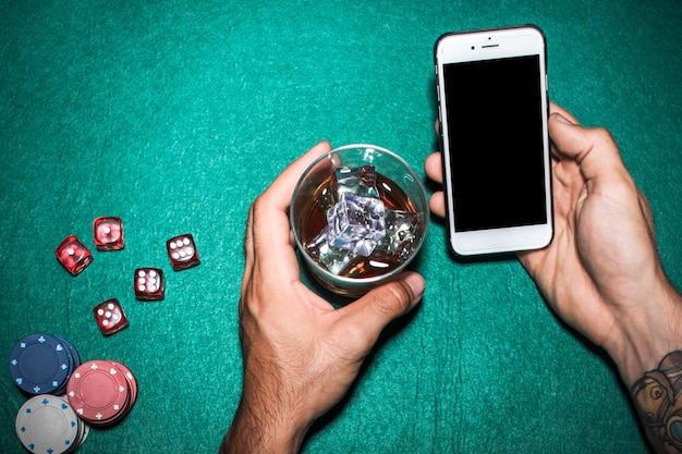 Free photo overhead view of man's hand holding cellphone and whisky glass over the poker table