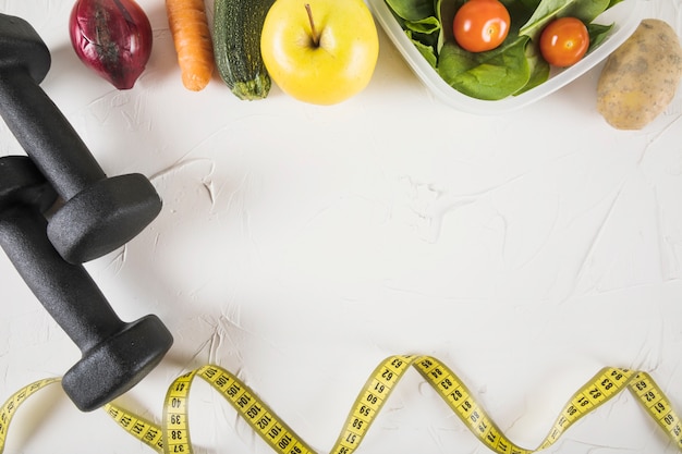 Overhead view of measuring tape; food and dumbbell on white backdrop