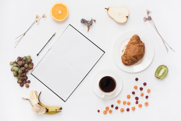 Free photo an overhead view of notebook; pen with croissant and healthy fruits isolated on white background