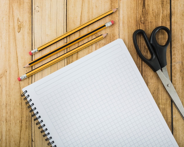 Overhead view of notepad; scissors and pencils on wooden plank