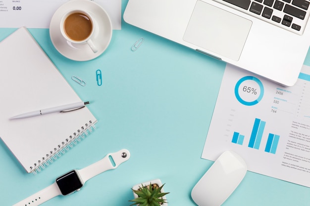 An overhead view of office desk with stationeries,laptop,mouse and smart watch on blue backdrop