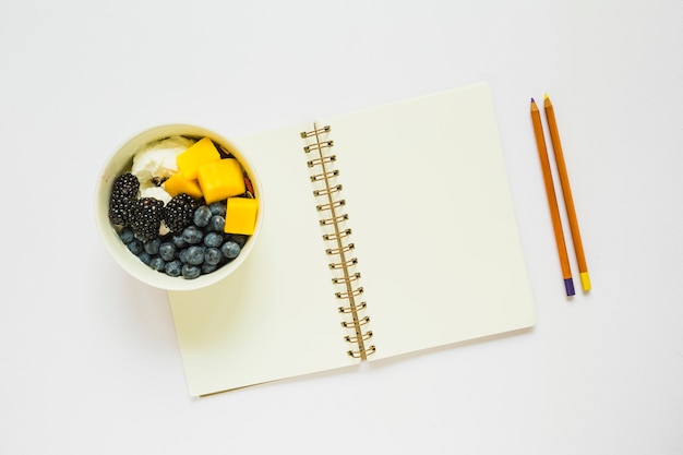 Free photo an overhead view of pencils and blank spiral notebook with healthy fruits in cup