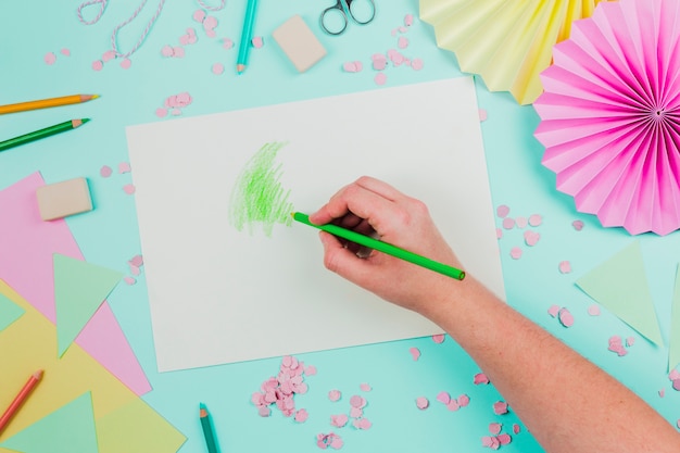 Free photo an overhead view of a person drawing with green pencil on white paper over the teal background