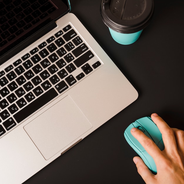 Free Photo an overhead view of person's hand using mouse with laptop and disposable coffee cup on black background