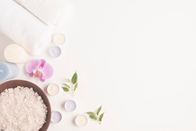 Overhead view of sea salt in clay bowl with orchid, candles and rolled up towel on white backdrop