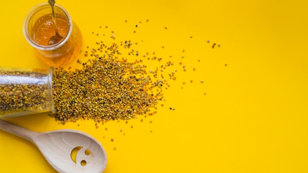 Free photo an overhead view of spilled bee pollens; honey pot and smiley wooden spoon on yellow backdrop